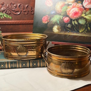 May include: Two oval brass planters with polished metallic finishes, featuring embossed scallop shell motifs on the sides and small side handles. The planters have a hammered texture around the upper rim and a smooth base, showcasing a warm golden tone.