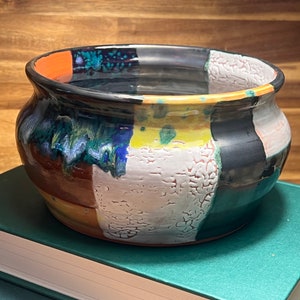 May include: A ceramic bowl with a colorful, geometric design. The bowl is glazed in shades of black, white, orange, yellow, and green. It has a textured surface and a rounded shape.