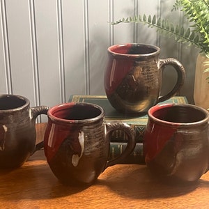 May include: Four ceramic mugs with a black and red glaze. The mugs have a rounded shape and a handle. The mugs are sitting on a wooden table.