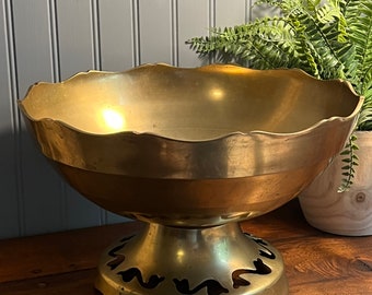 May include: A large, gold-toned brass bowl with a scalloped rim and a decorative base. The bowl is on a wooden surface.