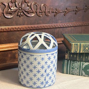 May include: A white ceramic jar with a blue and white star pattern. The jar has a lattice-like lid and is sitting on a table with a stack of books.