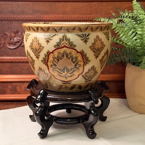 May include: A round ceramic bowl featuring a cream-colored base with a repeating pattern of black and gold botanical motifs and red accents. The bowl rests on a dark brown, carved wooden stand with four curved legs and a circular base.