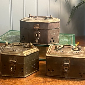May include: Three antique brass boxes with intricate designs and latches. The boxes have a hexagonal shape and are decorated with small holes. The boxes are sitting on a wooden surface.