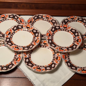 May include: A set of seven round ceramic plates featuring a cream-colored center and a decorative border with a repeating pattern of dark brown, orange, and white floral motifs. Each plate has a scalloped edge and a smooth, glazed finish.