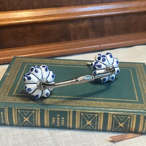 May include: A pair of blue and white ceramic drawer pulls with a silver metal connecting bar. The pulls are shaped like a flower with a raised center and are mounted on a book titled "The Razor's Edge" by W. Somerset Maugham.