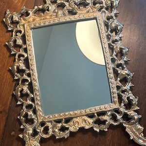 May include: A gold-toned ornate metal mirror with a decorative frame and three legs. The mirror has a rectangular shape and a clear glass surface.