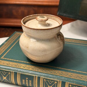 May include: A small, round, cream-colored ceramic jar with a matching lid. The pottery features a speckled glaze with brown accents around the rim and a small, circular decorative detail on the side. The surface has a smooth, glossy finish.