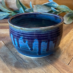 May include: A small, blue and brown ceramic bowl with a drip glaze design. The bowl is sitting on a wooden surface.