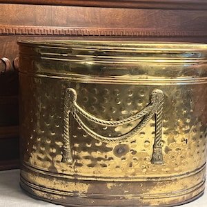May include: A large, oval-shaped brass planter with a hammered finish. The planter has a decorative rope design with tassels on each side.