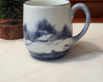 Puede incluir: Una taza de cerámica blanca con un paisaje invernal pintado a mano en azul que muestra una pequeña cabaña, suelo nevado y árboles desnudos. La taza tiene un asa curva y un borde azulado, con una firma visible cerca de la base del diseño.