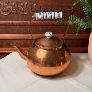 May include: A copper tea kettle with a rounded body and a short spout. The lid features a small white ceramic knob with blue floral accents. The arched metal handle includes a white ceramic grip decorated with repeating blue painted patterns.