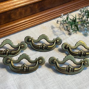 May include: Five antique brass cabinet pulls with a decorative, ornate design. The pulls have a curved shape with a center loop and are in excellent condition.