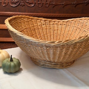 May include: A large, oval-shaped woven wicker basket with a natural light brown finish, featuring a wide, flared rim and a sturdy pedestal base. Two small decorative pumpkins, one cream and one sage green, sit beside the basket.