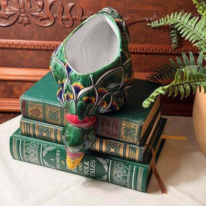 May include: A ceramic duck-shaped planter featuring a glossy green, white, and yellow glaze with a red neck band. The duck has a white interior and is positioned with its head facing downward, resting on a stack of three green hardcover books.