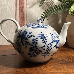 May include: A white teapot with a blue floral design. The teapot has a round body and a spout on the right side. The handle is on the left side. The teapot is sitting on a wooden surface.