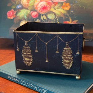 May include: A black metal planter with gold accents. The planter features a decorative design of gold pinecones and hanging ornaments. The planter is rectangular with small gold feet and handles. The planter is sitting on a book.