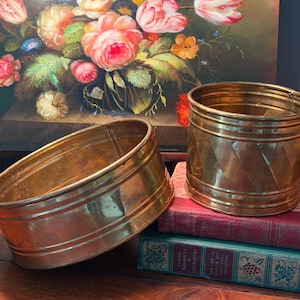May include: Two antique brass planters, one oval and one cylindrical, sit on a wooden surface atop a stack of books. The oval planter has horizontal bands, while the cylindrical one has a diamond pattern. A floral painting is in the background.