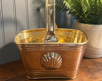 May include: A gold-colored metal basket with a shell design on the front and a handle. The basket is sitting on a wooden surface.