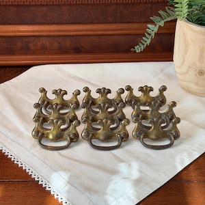 May include: A set of six brass drawer pulls featuring an ornate, symmetrical design with a curved handle and a decorative backplate. Each piece has a weathered, antique finish with visible patina, showcasing a detailed scrollwork pattern.
