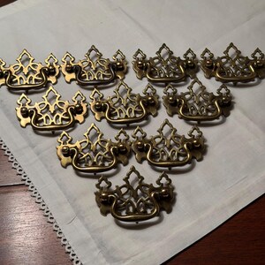 May include: A collection of ten brass drawer pulls arranged in a triangular formation. Each pull features an ornate, openwork backplate with a crown-like silhouette and a curved, swinging handle attached by two small metal posts on each side.