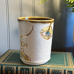 May include: White ceramic mug with a brown rim and a gold and blue painted design of a face. The mug is sitting on a green book with gold lettering that reads "The Razor's Edge".