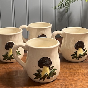 May include: Set of four white ceramic mugs with brown mushroom designs. Each mug has a different mushroom design with green leaves and a yellow stem.