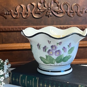 May include: White ceramic bowl with a black rim and a scalloped edge. The bowl is decorated with purple flowers and green leaves. The bowl is sitting on a black book with gold lettering that reads "The Confessions of Saint Augustine".