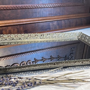 May include: A gold-toned metal tray with a mirrored surface and an ornate, decorative border. The tray is sitting on a white surface with dried lavender flowers.