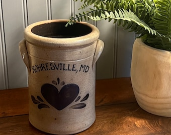 May include: A cream-colored stoneware crock with black painted details. The crock has two handles and a heart design with a black painted inscription that reads "Rohresville, MD".