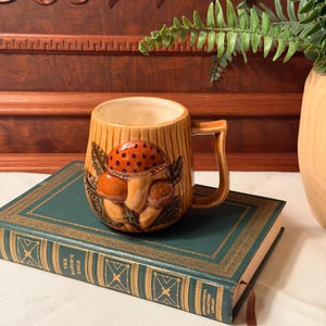 May include: A tan ceramic mug featuring a raised, three-dimensional relief of brown and orange mushrooms with green leaves. The mug has a textured, wood-grain pattern on its exterior, a smooth cream-colored interior, and a square-shaped handle.