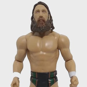 May include: A wrestling action figure with a tan muscular physique, long brown hair, and a beard. The figure wears black and green trunks, black and green knee-high boots, and white wristbands. The figure is posed with arms at its sides.
