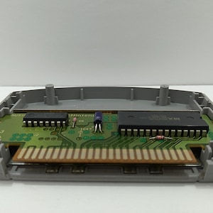 May include: An open Nintendo 64 game cartridge, revealing the internal circuit board. The board is green with various electronic components, including chips, resistors, and a connector. The cartridge casing is gray.