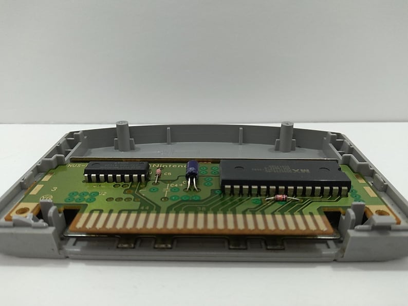 May include: An open Nintendo 64 game cartridge, revealing the internal circuit board. The board is green with various electronic components, including chips, resistors, and a connector. The cartridge casing is gray.