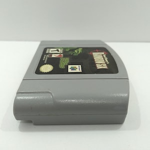 May include: A gray Nintendo 64 video game cartridge for Rainbow Six. The label features the game title, a green graphic, and the Nintendo logo. The cartridge is made of plastic and has a rectangular shape.