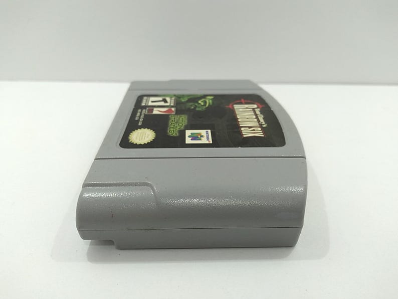 May include: A gray Nintendo 64 video game cartridge for Rainbow Six. The label features the game title, a green graphic, and the Nintendo logo. The cartridge is made of plastic and has a rectangular shape.
