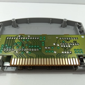 May include: A gray plastic video game cartridge shell with a green circuit board. The circuit board has gold-colored pins and various electronic components. The cartridge is open, revealing the internal components.