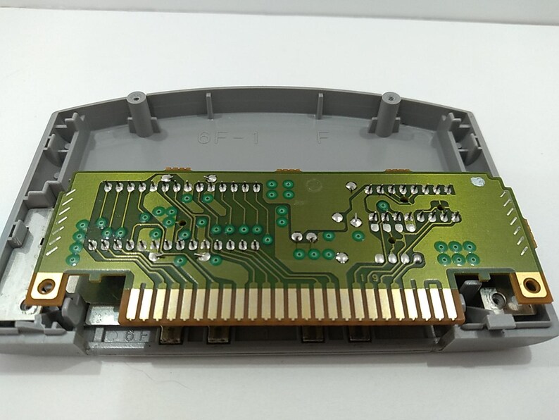 May include: A gray plastic video game cartridge shell with a green circuit board. The circuit board has gold-colored pins and various electronic components. The cartridge is open, revealing the internal components.