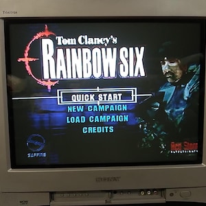 May include: A silver Sony Trinitron television displays the menu screen of the video game "Tom Clancy's Rainbow Six." The game's title is in white letters with a red and orange target logo. Menu options include Quick Start, New Campaign, Load Campaign, and Credits.