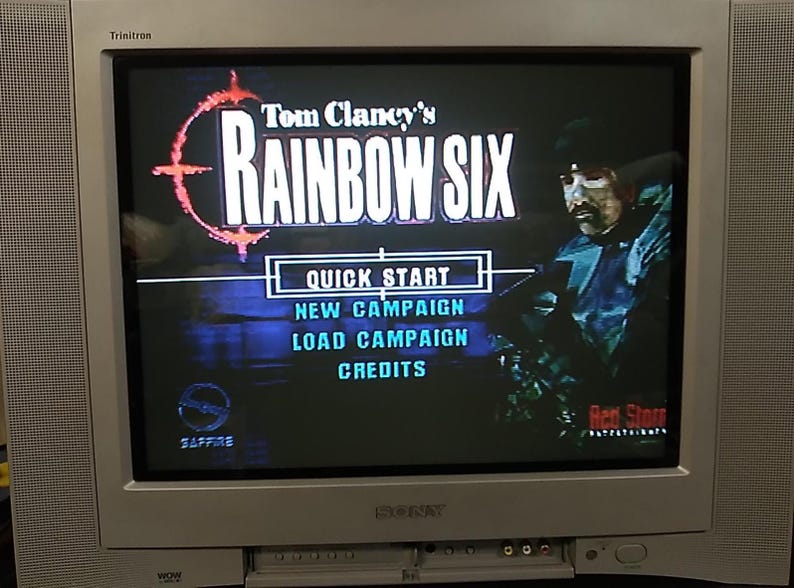 May include: A silver Sony Trinitron television displays the menu screen of the video game "Tom Clancy's Rainbow Six." The game's title is in white letters with a red and orange target logo. Menu options include Quick Start, New Campaign, Load Campaign, and Credits.