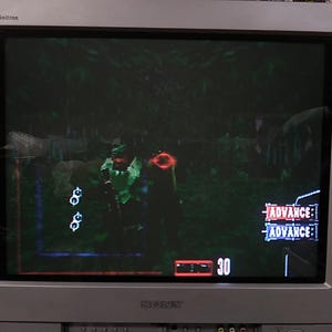 May include: A silver Sony Trinitron television displays a video game scene. The game features a green-clad character aiming a weapon in a forest setting. The screen shows health and ammo indicators, along with the word "ADVANCE".