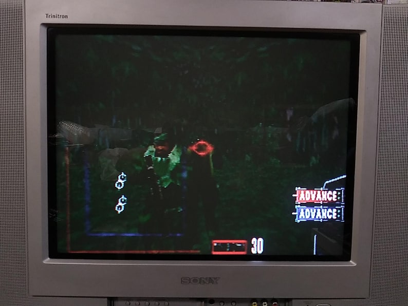 May include: A silver Sony Trinitron television displays a video game scene. The game features a green-clad character aiming a weapon in a forest setting. The screen shows health and ammo indicators, along with the word "ADVANCE".