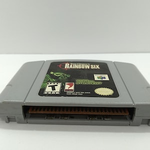 May include: Gray Nintendo 64 video game cartridge for Tom Clancy's Rainbow Six. The label features the game title, a green and black graphic, and the Nintendo logo. The cartridge is rated T for Teen.