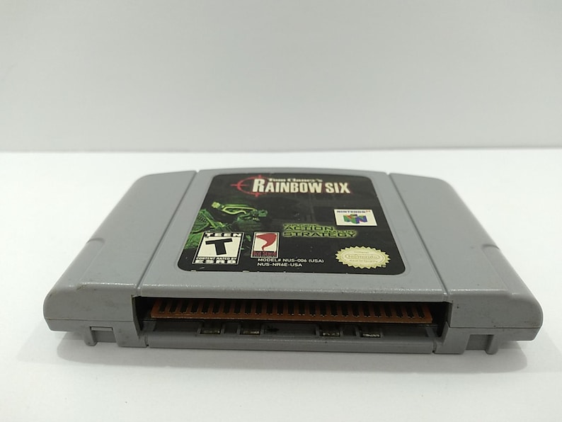 May include: Gray Nintendo 64 video game cartridge for Tom Clancy's Rainbow Six. The label features the game title, a green and black graphic, and the Nintendo logo. The cartridge is rated T for Teen.