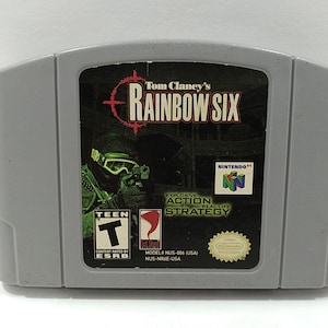 May include: A gray Nintendo 64 game cartridge for "Tom Clancy's Rainbow Six." The game's label features a soldier in tactical gear, the game's title, and the ESRB Teen rating. The game is an explosive action real-life strategy game.