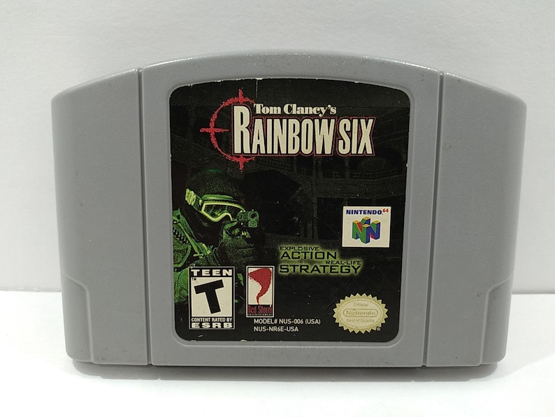 May include: A gray Nintendo 64 game cartridge for "Tom Clancy's Rainbow Six." The game's label features a soldier in tactical gear, the game's title, and the ESRB Teen rating. The game is an explosive action real-life strategy game.