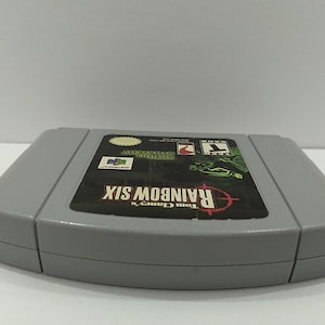 May include: A gray Nintendo 64 video game cartridge for Tom Clancy's Rainbow Six. The label features the game's title in red, a target graphic, and various logos. The cartridge is in good condition.