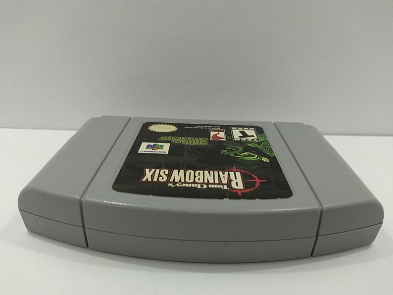 May include: A gray Nintendo 64 video game cartridge for Tom Clancy's Rainbow Six. The label features the game's title in red, a target graphic, and various logos. The cartridge is in good condition.