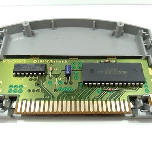 May include: An open Nintendo 64 game cartridge, showcasing the internal circuit board. The board is green with various electronic components, including chips and connectors. The cartridge casing is gray, and the board has the text "1996 Nintendo" and "NUS-01A-02".