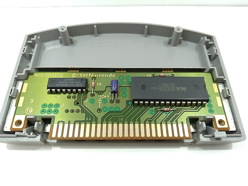 May include: An open Nintendo 64 game cartridge, showcasing the internal circuit board. The board is green with various electronic components, including chips and connectors. The cartridge casing is gray, and the board has the text "1996 Nintendo" and "NUS-01A-02".