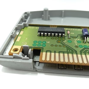 May include: Close-up of a Nintendo 64 game cartridge interior. The green circuit board features various electronic components, including chips, resistors, and capacitors. The cartridge is partially encased in a gray plastic shell.